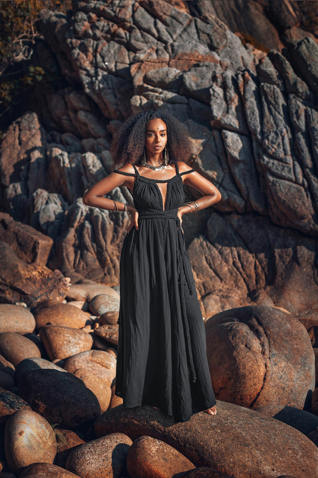 A woman stands confidently amidst rugged terrain, dressed in an elegant Black Greek Goddess Prom Boho Evening Dress from AYA Sacred Wear. Her hands rest on her hips, and her voluminous hair is beautifully highlighted by the warm, natural light against a backdrop of large stones and jagged rocks.