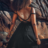 A woman with curly hair stands on rocky terrain, wearing the Black Greek Goddess Prom Boho Evening Dress by AYA Sacred Wear, which features a sleeveless design and a low back with criss-cross straps. Her skin glistens in the sunlight.