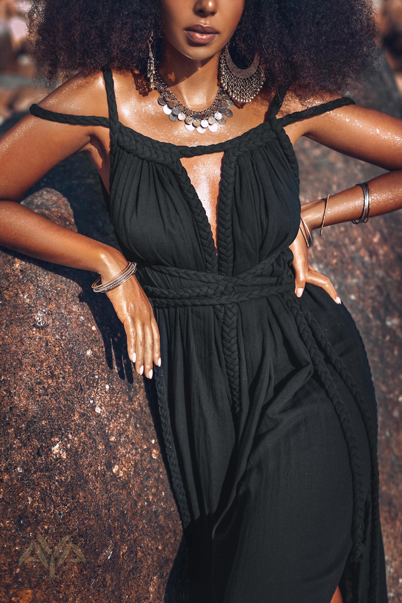 A woman with curly hair stands by a rock, wearing the Black Greek Goddess Prom Boho Evening Dress from AYA Sacred Wear. Her organic cotton ensemble is complemented by silver jewelry, including large hoop earrings, a necklace, and bracelets. The sun highlights her skin and the texture of her outfit beautifully.