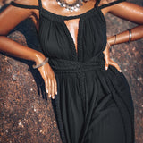 A woman with curly hair stands by a rock, wearing the Black Greek Goddess Prom Boho Evening Dress from AYA Sacred Wear. Her organic cotton ensemble is complemented by silver jewelry, including large hoop earrings, a necklace, and bracelets. The sun highlights her skin and the texture of her outfit beautifully.