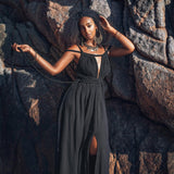 Wearing an elegant Black Greek Goddess Prom Boho Evening Dress by AYA Sacred Wear, a woman poses against large, textured rocks. Her curly hair complements her hoop earrings and bracelets as she gracefully holds one arm high while the other rests on the rock. Warm sunlight bathes the scene, highlighting her effortless style.
