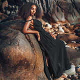 A person in a Black Greek Goddess Prom Boho Evening Dress by AYA Sacred Wear is leaning against a large rock surrounded by smaller ones, creating a serene outdoor ambiance in a rocky setting. The person's voluminous curls perfectly enhance the natural vibe of the scene.