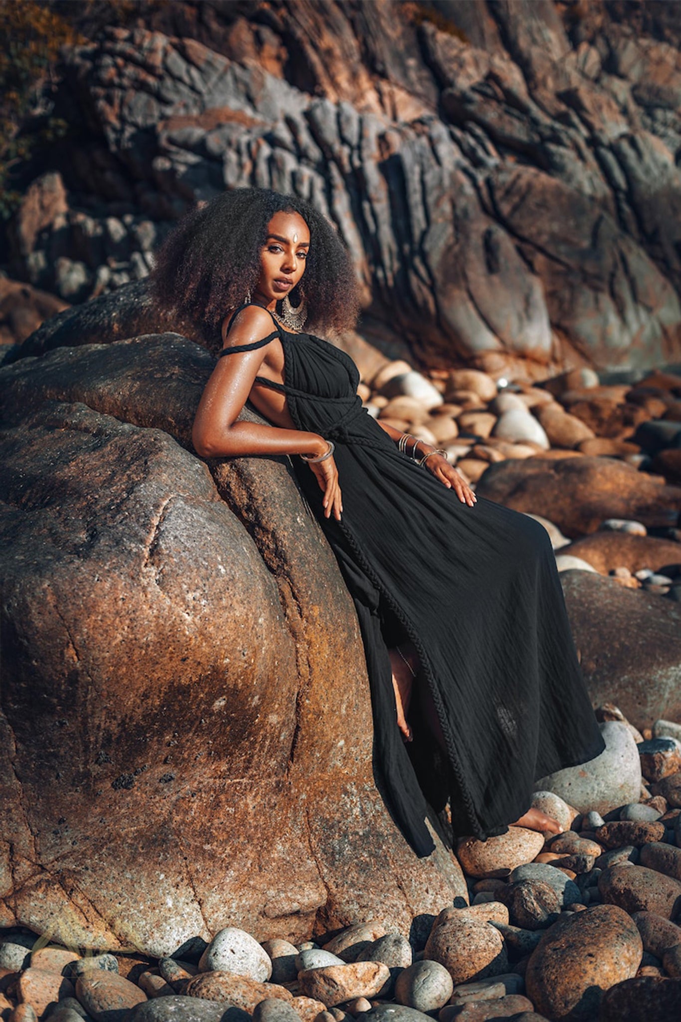 A person in a Black Greek Goddess Prom Boho Evening Dress by AYA Sacred Wear is leaning against a large rock surrounded by smaller ones, creating a serene outdoor ambiance in a rocky setting. The person's voluminous curls perfectly enhance the natural vibe of the scene.