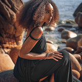 A woman in an AYA Sacred Wear Black Greek Goddess Prom Boho Evening Dress sits on a rocky seaside, with the ocean in the background. Her curly hair and jewelry add to her contemplative and serene presence under the bright sky.