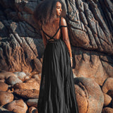 A woman with curly hair stands on rocky terrain, elegantly dressed in the Black Greek Goddess Prom Boho Evening Dress by AYA Sacred Wear. The long, flowing gown with thin straps is crafted from organic cotton. Her arms are adorned with golden bracelets, while large, jagged rocks bask in the warm sunlight behind her.
