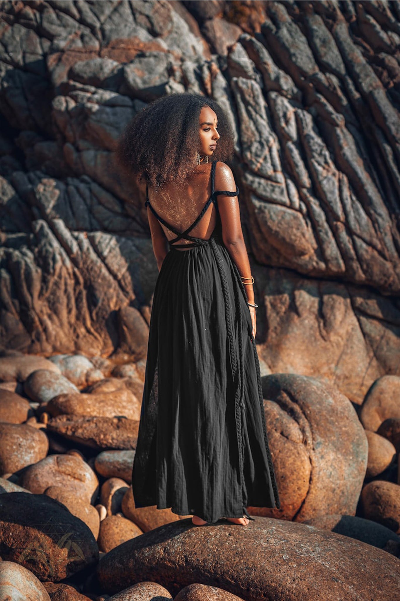 A woman with curly hair stands on rocky terrain, elegantly dressed in the Black Greek Goddess Prom Boho Evening Dress by AYA Sacred Wear. The long, flowing gown with thin straps is crafted from organic cotton. Her arms are adorned with golden bracelets, while large, jagged rocks bask in the warm sunlight behind her.