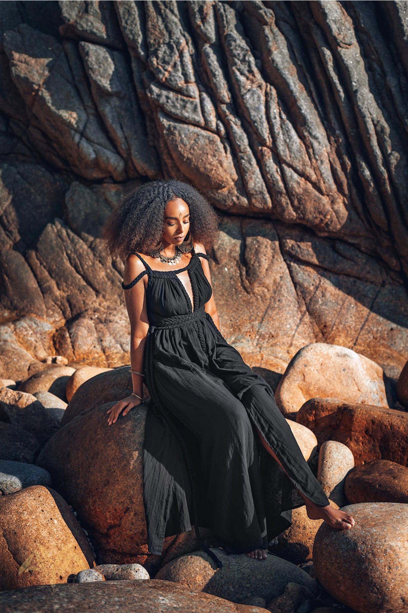 A woman in an elegant Black Greek Goddess Prom Boho Evening Dress from AYA Sacred Wear sits gracefully on large rocks. She is surrounded by a rugged, sunlit rock formation, creating a serene and contemplative atmosphere.