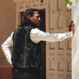In a relaxed pose, a person with long hair leans against a wall outdoors. They are wearing an AYA Sacred Wear Black Hand Embroidered Vest for Men over a white shirt and black pants. The background features an elegant, ornate wall adorned with a hanging lantern and botanically dyed flowers seen above.