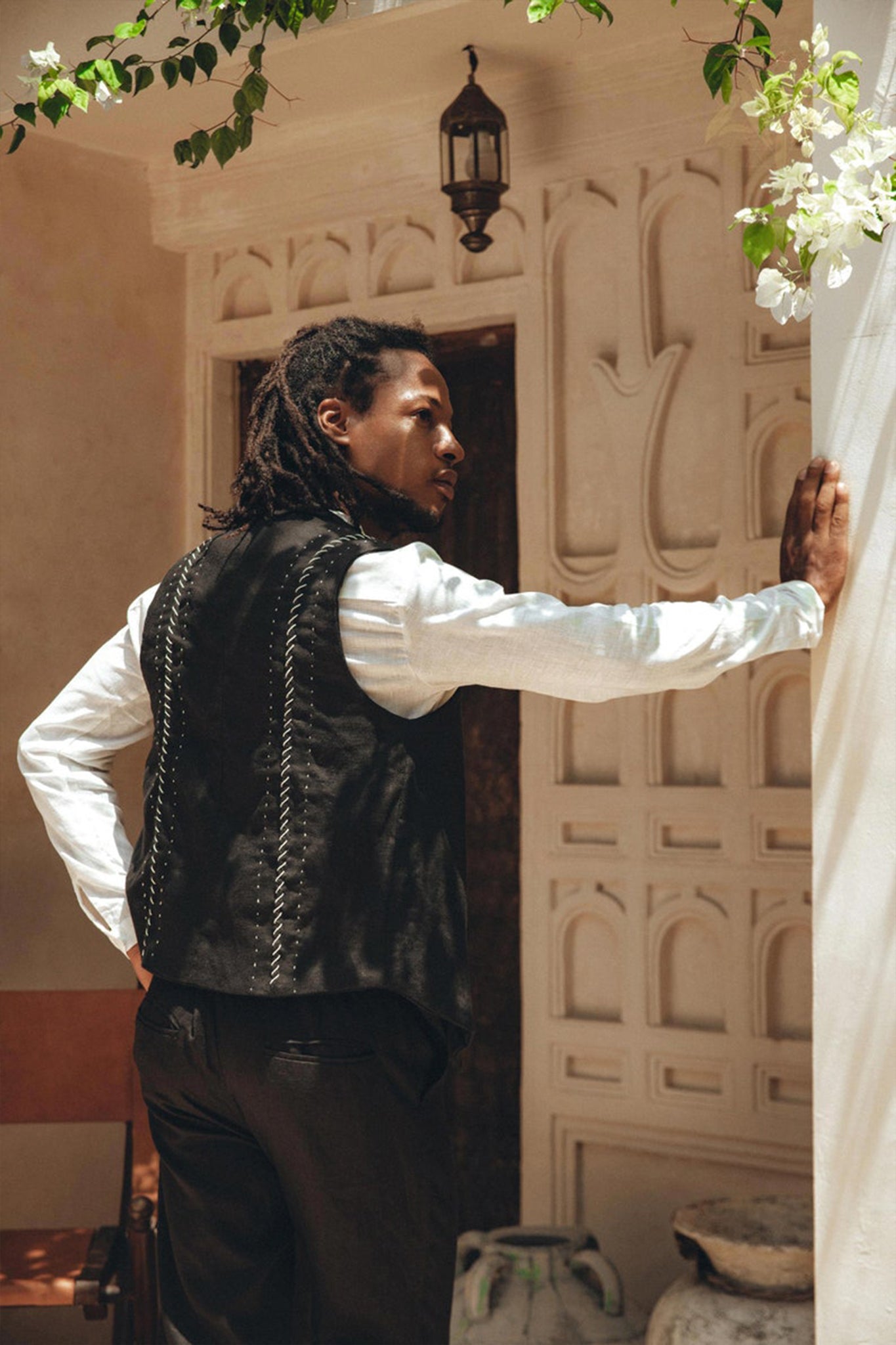 In a relaxed pose, a person with long hair leans against a wall outdoors. They are wearing an AYA Sacred Wear Black Hand Embroidered Vest for Men over a white shirt and black pants. The background features an elegant, ornate wall adorned with a hanging lantern and botanically dyed flowers seen above.