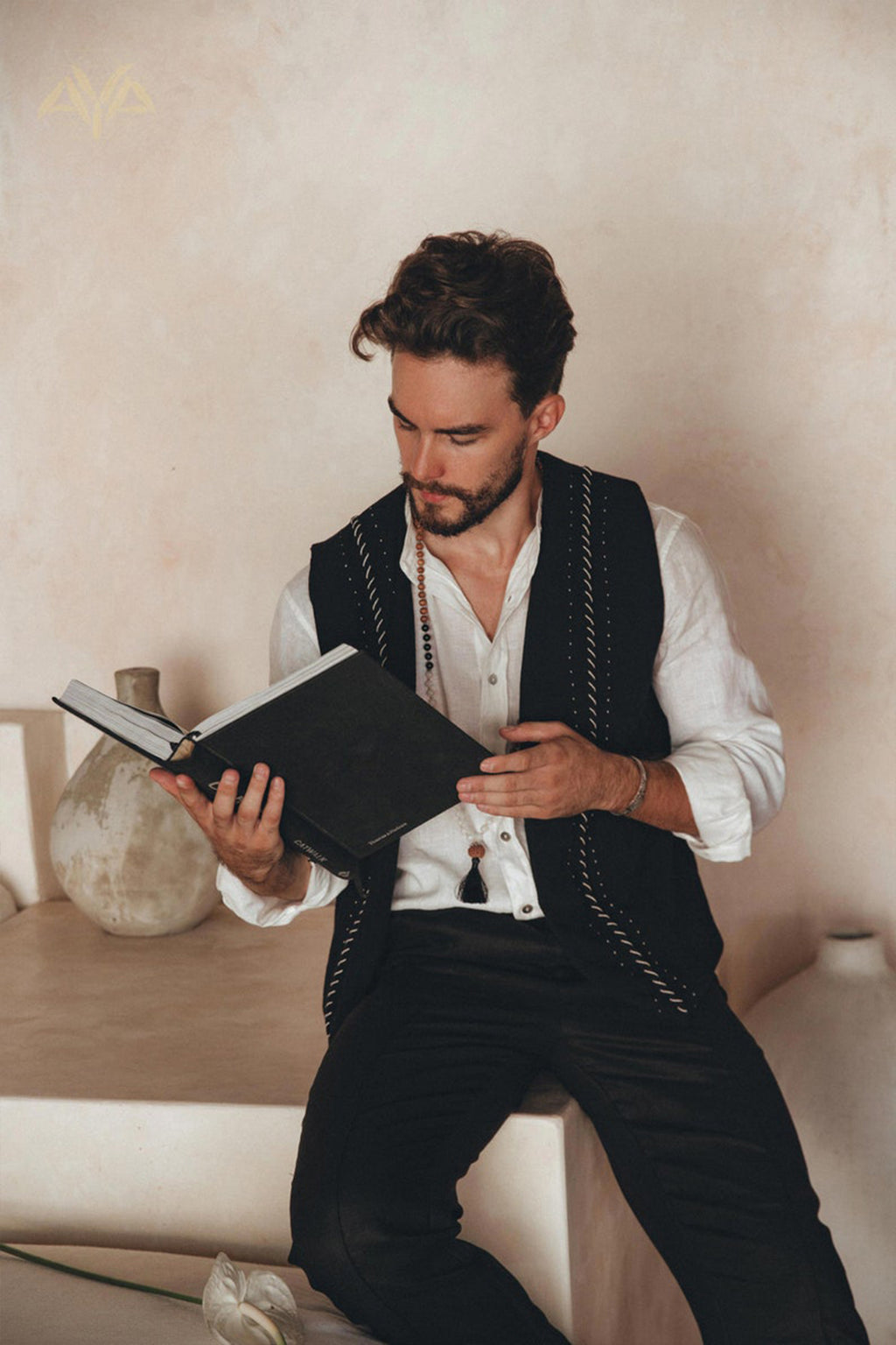 A man with dark hair and a beard is sitting on a ledge, engrossed in reading a book. He wears the Black Hand Embroidered Vest for Men from AYA Sacred Wear over his white shirt. Ceramic vases add elegance to the background against the textured beige wall.