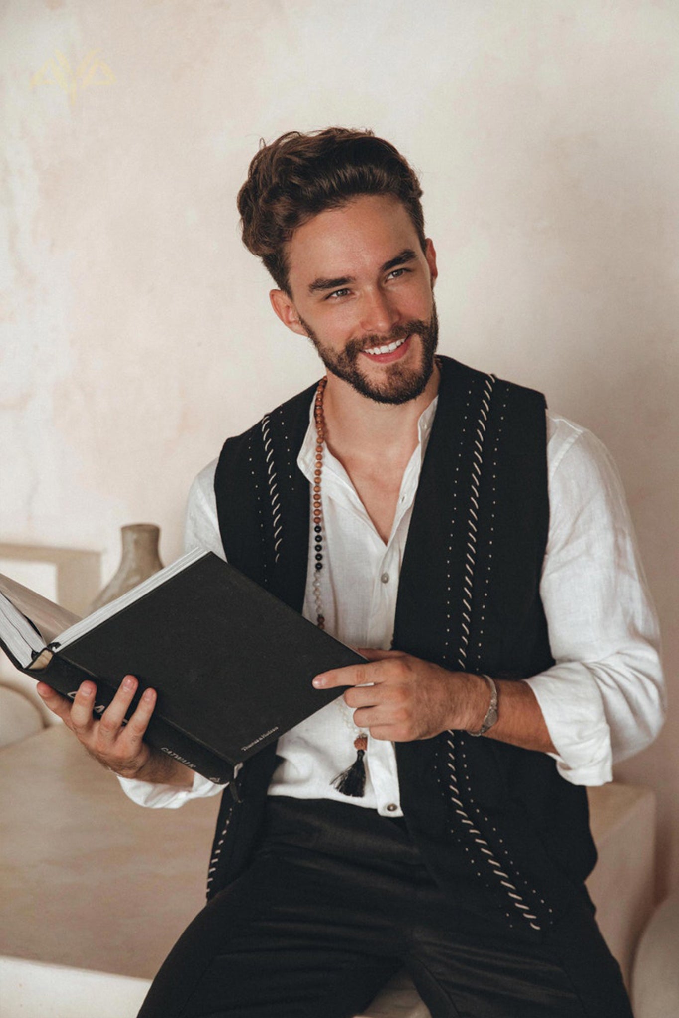 A man with a beard, wearing a white shirt and a Black Hand Embroidered Vest from AYA Sacred Wear, is sitting indoors and smiling while holding an open black book.