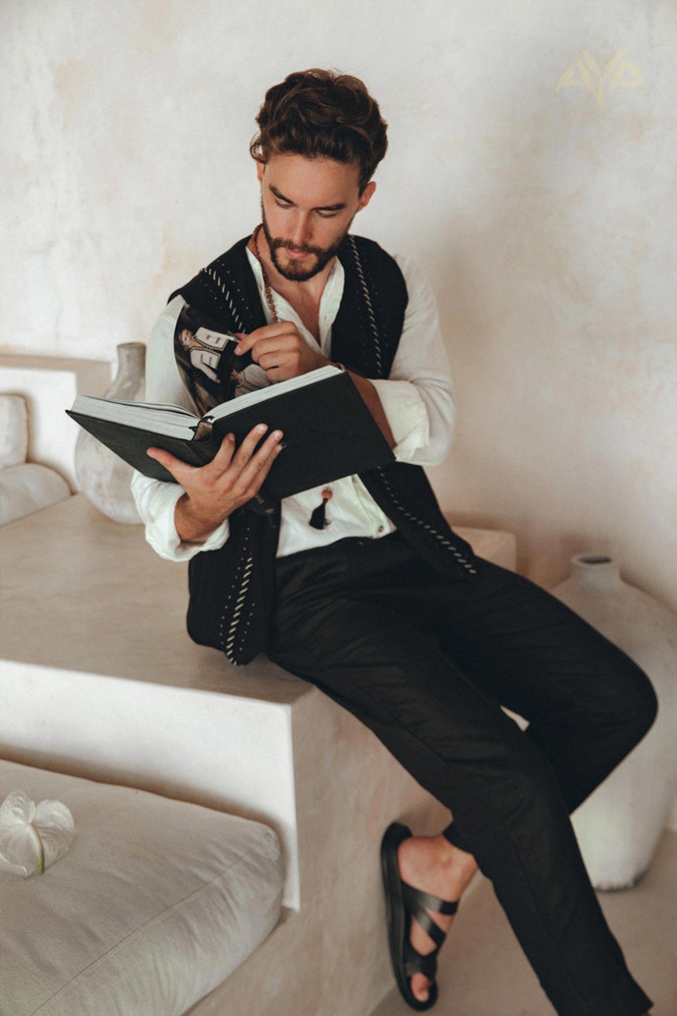 A man sits on a ledge, dressed in the Black Hand Embroidered Vest for Men by AYA Sacred Wear, layered over a white shirt and black pants. He is engrossed in reading a book, with a pen in hand. The minimalist background features neutral tones and decorative pottery.