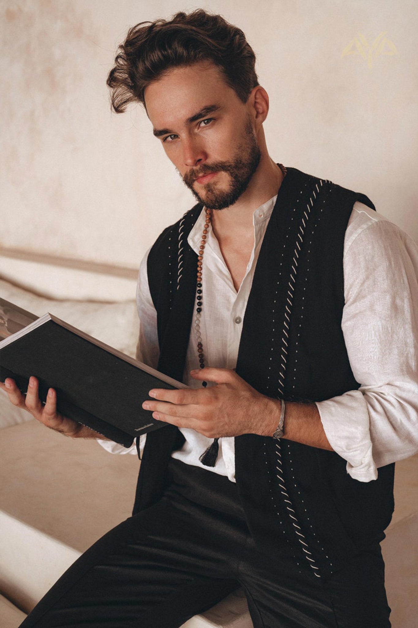 A man with a beard sits on a staircase, cradling a book. His black hand-embroidered vest from AYA Sacred Wear, crafted from organic cotton, complements his white shirt against the backdrop of a minimalist, light-toned wall.