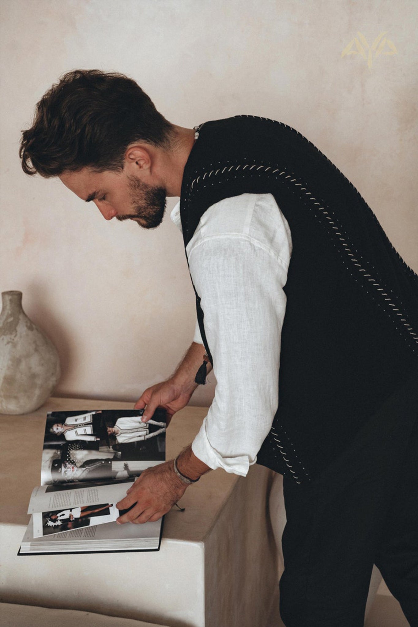 A person with short hair and a beard, wearing an AYA Sacred Wear Black Hand Embroidered Vest over a white shirt, is standing and flipping through a magazine. A beige vase sits on the table next to them against a neutral-toned wall.