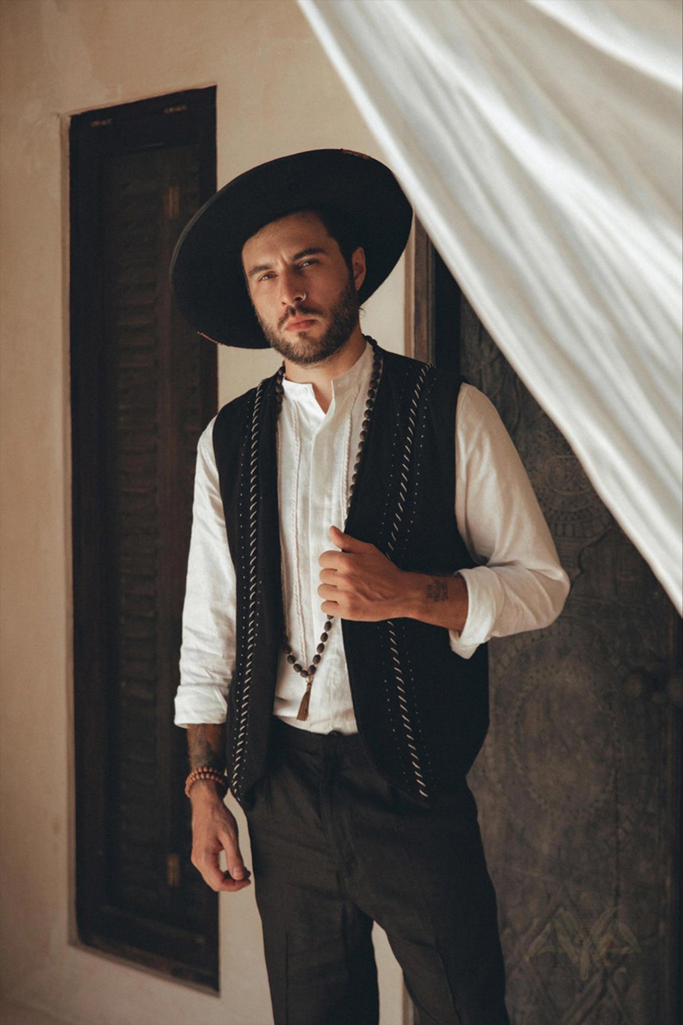 A person with a beard stands indoors wearing a wide-brimmed hat, a white shirt, and the Black Hand Embroidered Vest for Men by AYA Sacred Wear. They hold a string of beads and stand next to a draped curtain.