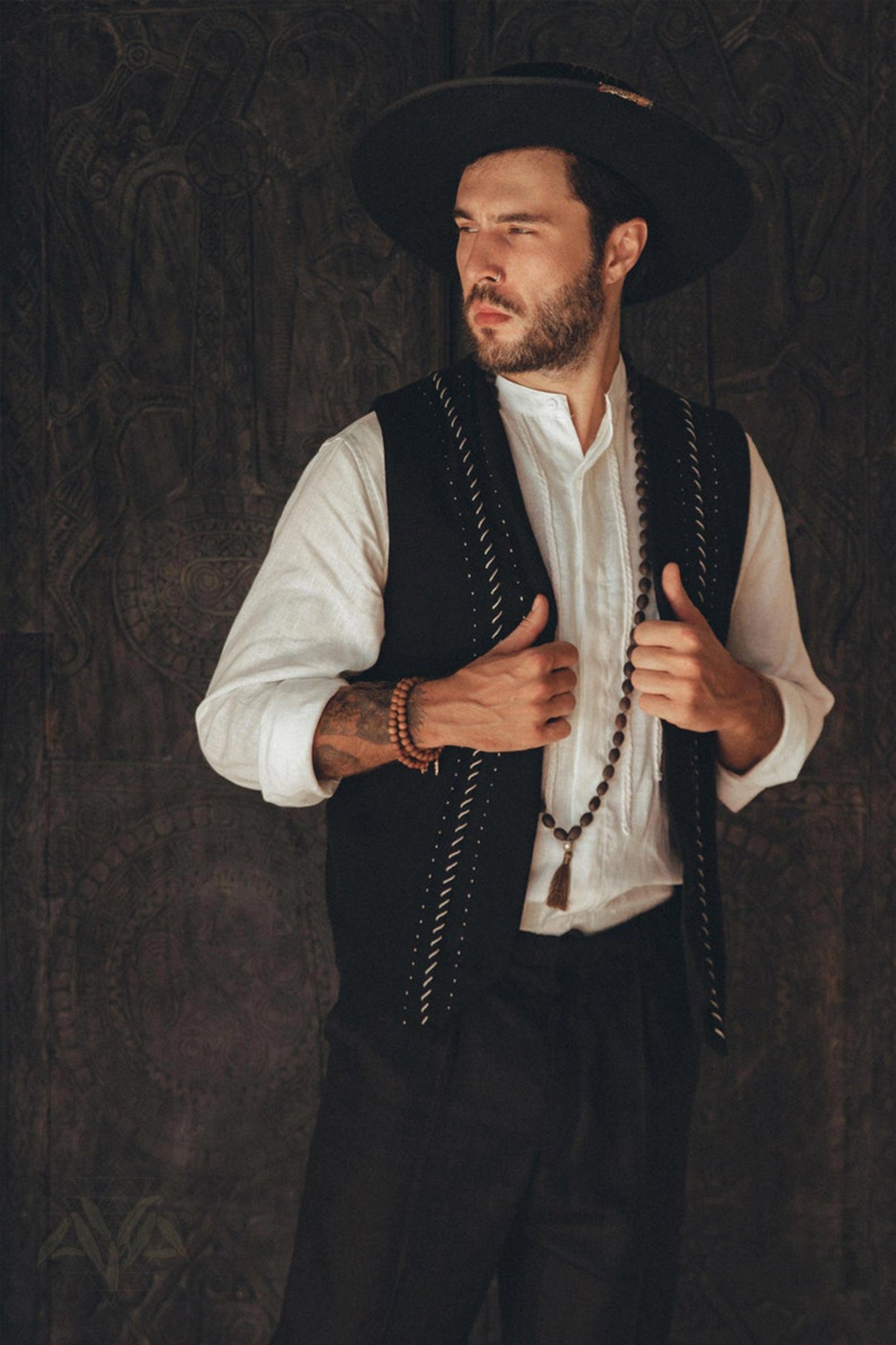 A bearded man dons a wide-brimmed hat, an AYA Sacred Wear Black Hand Embroidered Vest over a white shirt, and a long beaded necklace. He stands with a thoughtful expression against a dark, textured background, exuding an air of timeless elegance and natural style.