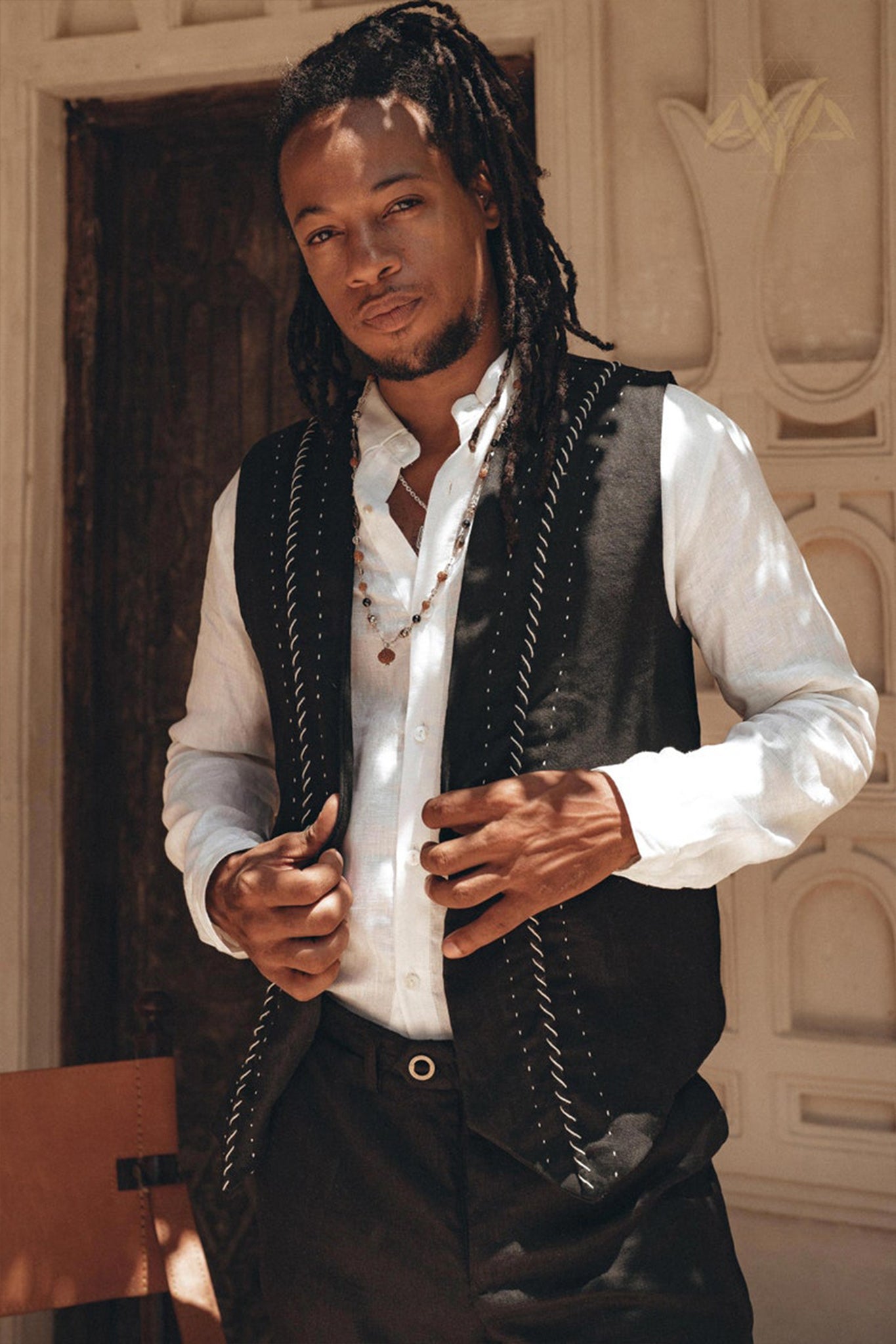 A person with long hair stands in front of a textured wall, wearing a white shirt and an AYA Sacred Wear Black Hand Embroidered Vest for Men that appears botanically dyed. The necklace adds flair to their relaxed expression as they adjust the handwoven vest. A wooden chair is partially visible beside them.