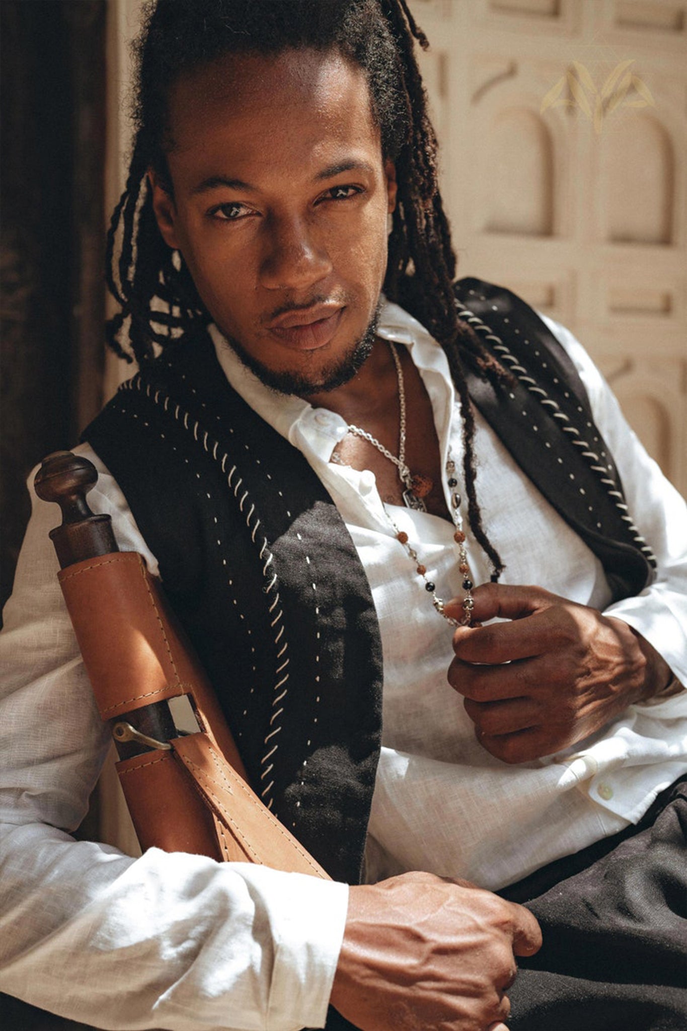 A person with long braided hair poses confidently, wearing a white shirt paired with the Black Hand Embroidered Vest for Men by AYA Sacred Wear. They have a leather strap across their chest and stand against a textured wall, exuding style with the artisanal touch of handwoven sophistication.