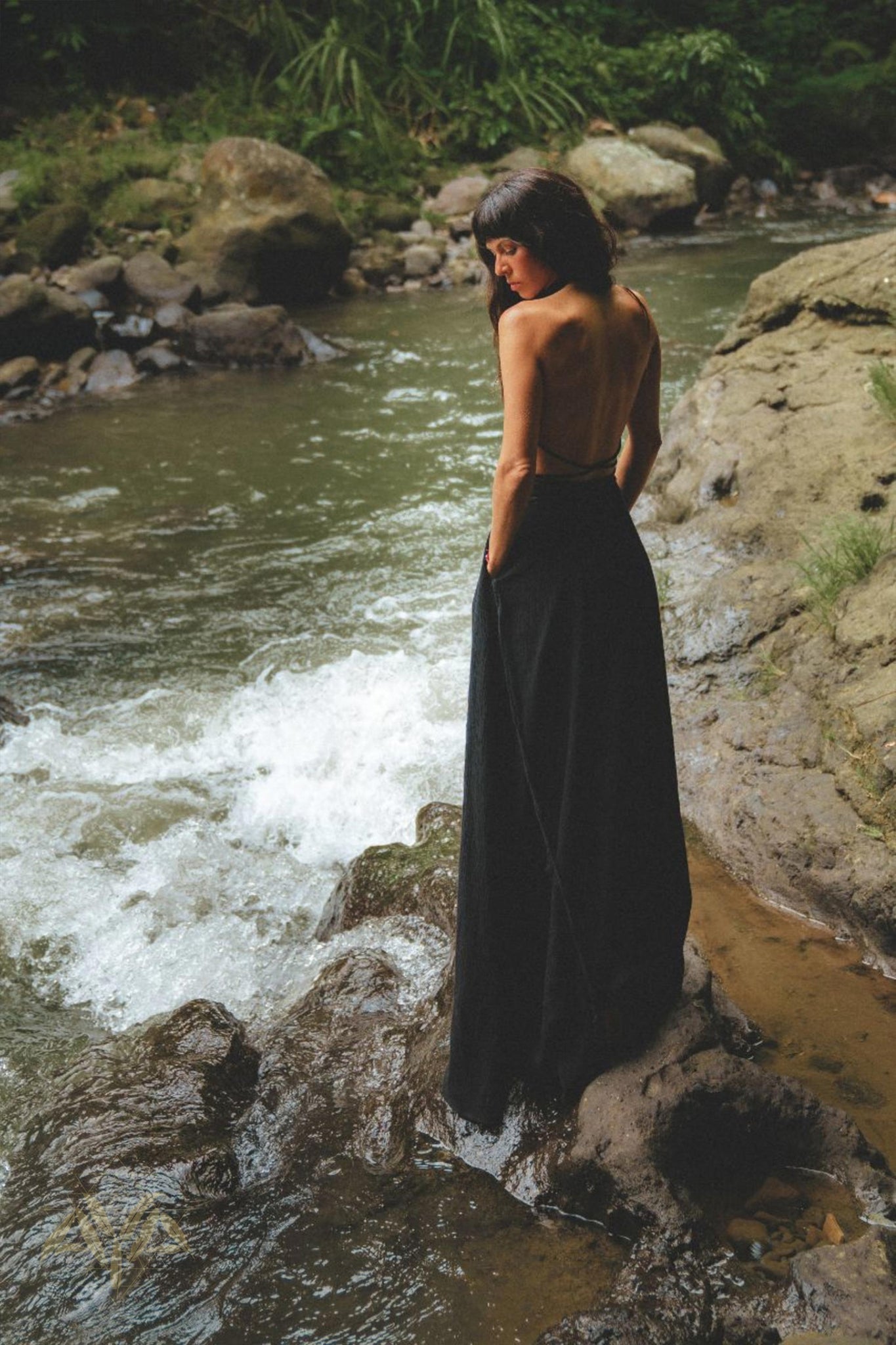 A person with long hair stands on a rocky riverbank, facing a flowing stream. They are wearing an elegant Black Harmonia Dress by AYA Sacred Wear, made from organic fabric. The dress, known for its adjustable and Gothic sexy style, adds to the enchanting look of the "Fairy of the River" scene. Lush green foliage surrounds them, creating a serene and natural atmosphere as they appear to be contemplating the water.