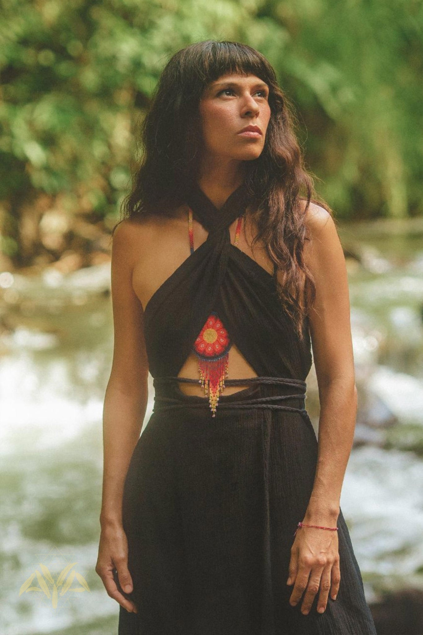 A woman with long, wavy hair stands outdoors with a serene forest and river in the background. She is wearing a black, halter-neck Black Harmonia Dress by AYA Sacred Wear, known for its gothic and sexy style as well as its fairy-like charm. The dress, made from organic fabric, is complemented by a beaded necklace featuring a red pendant. Her expression appears thoughtful as she gazes off into the distance.