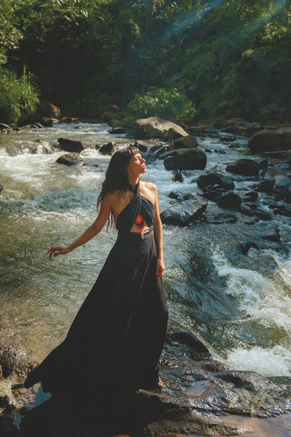 A woman stands on a rock in a flowing Black Harmonia Dress by the river. Sunlight filters through the trees, illuminating her face and the lush, green surroundings. The scenery, with its vibrant vegetation and flowing river, perfectly complements her dress—a blend of organic linen and cotton from AYA Sacred Wear's Gothic Sexy collection, also known as the Fairy of the River Dress.