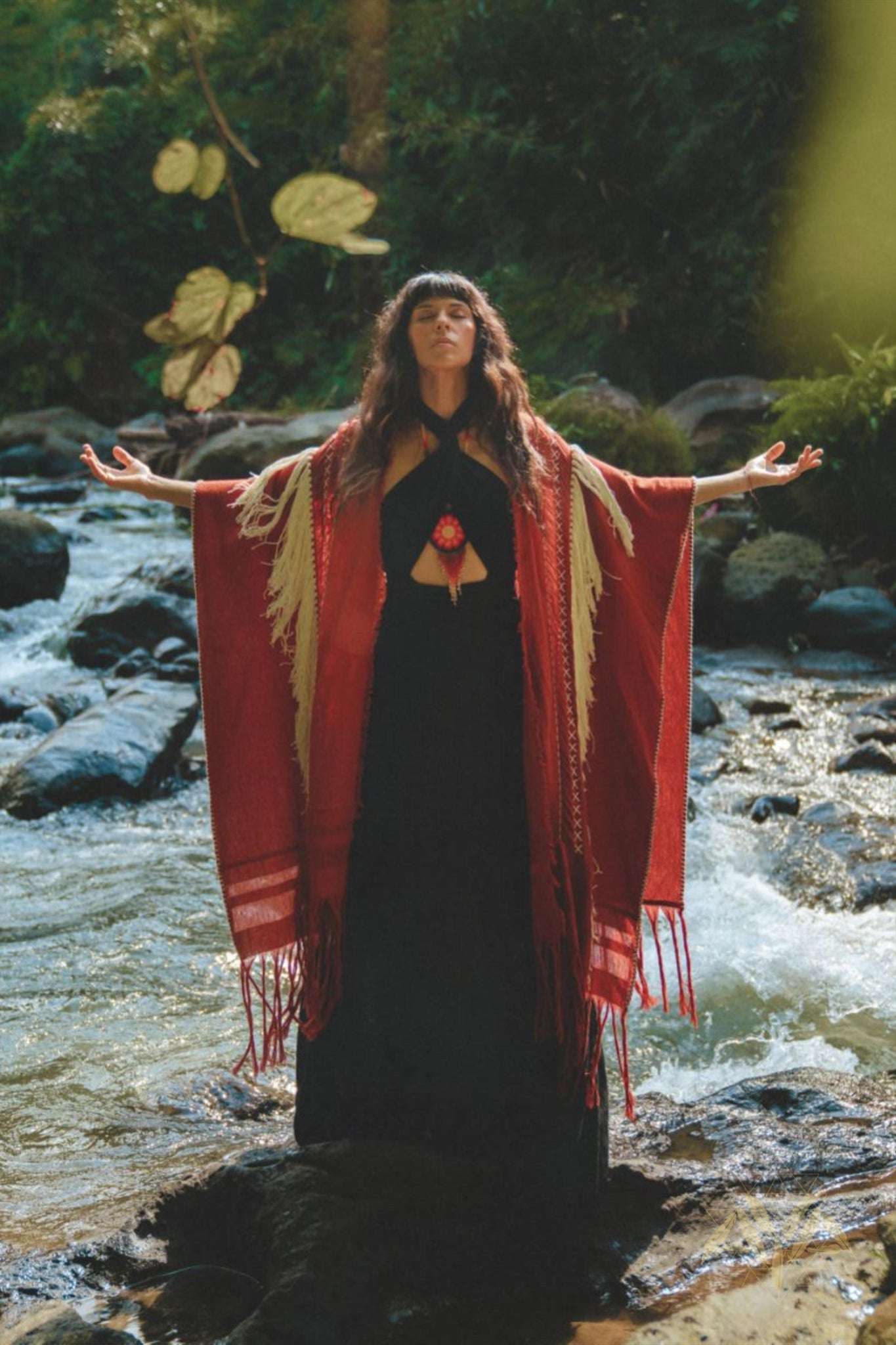 A woman stands on rocks in a stream, arms spread wide and looking upward. She is wearing the Black Harmonia Dress from AYA Sacred Wear, a Gothic sexy, adjustable dress also known as the Fairy of the River Dress. The dress is crafted from organic linen and cotton and is complemented by a red and beige shawl. The surrounding area is lush with greenery as sunlight filters through the trees.