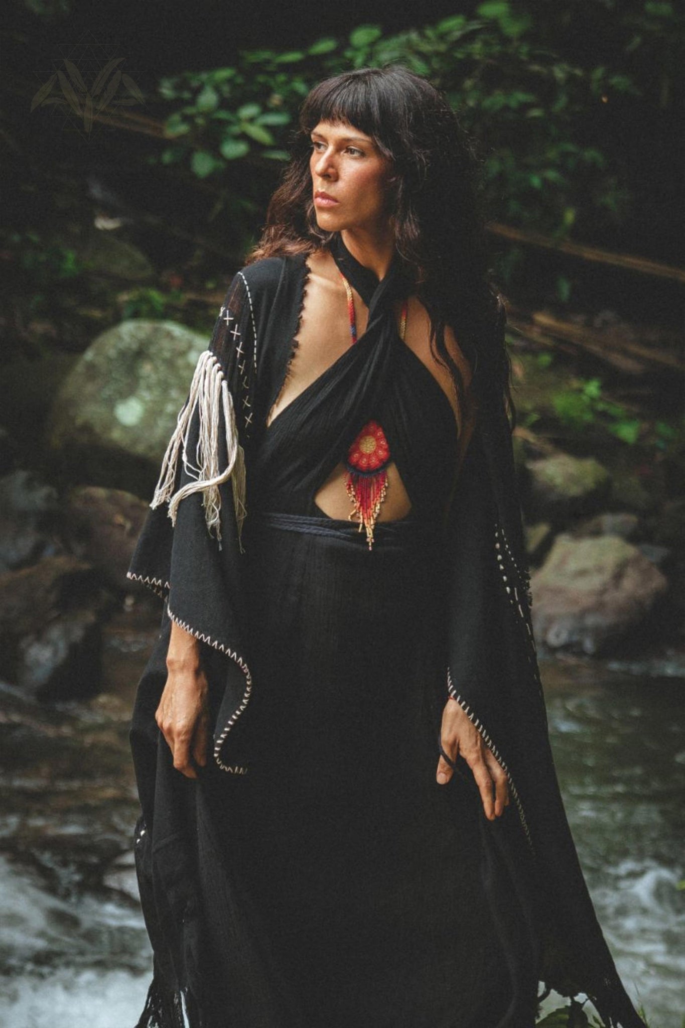 A woman stands outdoors near a stream and rocks, surrounded by greenery. She wears the Black Harmonia Dress by AYA Sacred Wear, which is an adjustable dress made from handwoven fabric with stitched details and a red adornment at the chest. Her dark hair is loose, and she gazes slightly to the side with a contemplative expression.