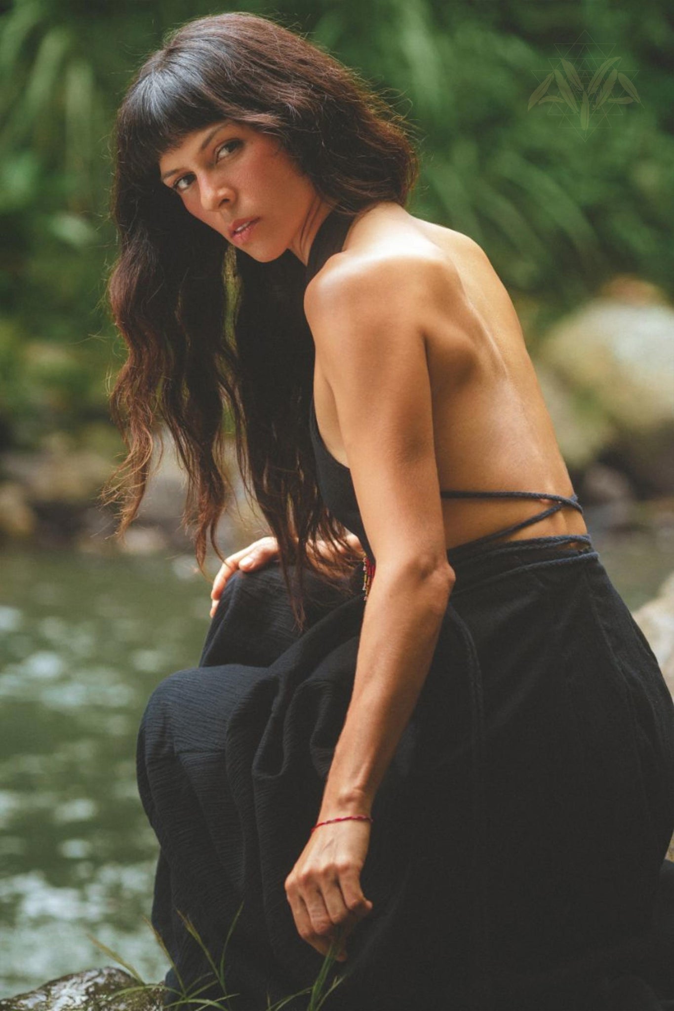 A woman with long dark hair poses by a serene river, wearing the elegant Black Harmonia Dress from AYA Sacred Wear. This adjustable Gothic sexy dress, also known as the Fairy of the River Dress, features a low, strappy back. She gazes over her shoulder towards the camera, surrounded by lush green foliage and rocks. The dress is crafted from organic linen and cotton and enhanced with botanical dyes.