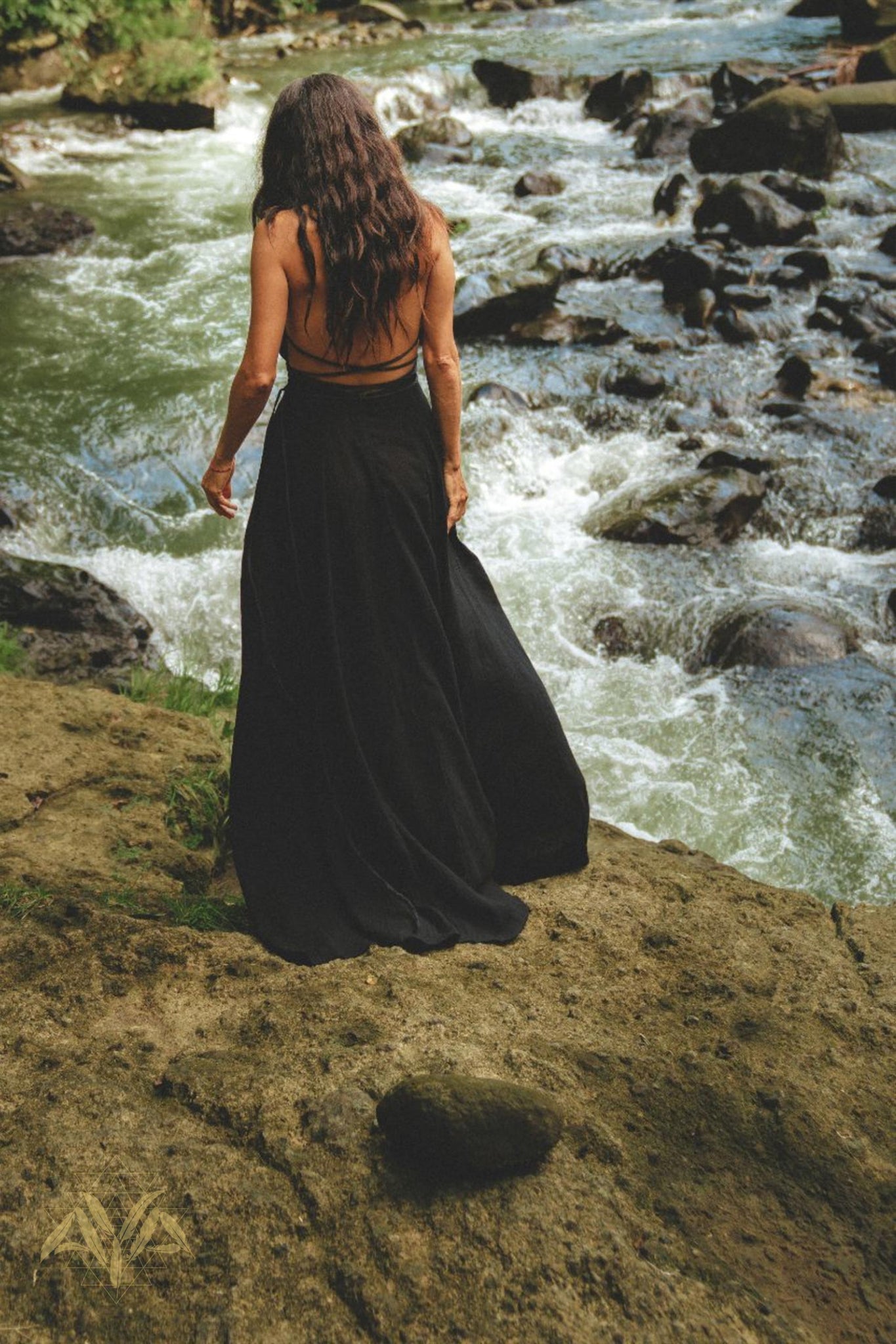 A woman with long, wavy hair, dressed in a flowing Black Harmonia Dress from AYA Sacred Wear—an adjustable Gothic sexy dress known as the Fairy of the River Dress and made from organic linen and cotton dyed with botanical dyes—stands on a rocky ledge overlooking a fast-moving stream surrounded by rocks and greenery. The scene exudes a sense of peace and natural beauty.