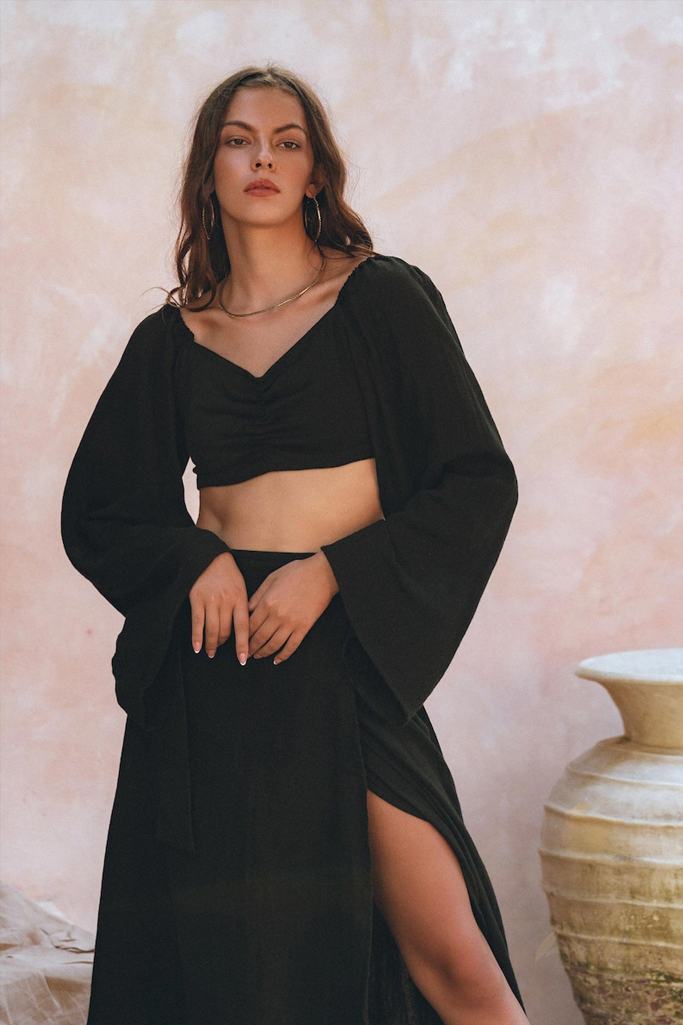 A person wearing the Black Summer Open Belly Top and Skirt by AYA Sacred Wear poses against a soft pink textured background. They have long hair and accessorize with hoop earrings. A large ceramic pot is partially visible on the right.