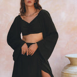 A person wearing the Black Summer Open Belly Top and Skirt by AYA Sacred Wear poses against a soft pink textured background. They have long hair and accessorize with hoop earrings. A large ceramic pot is partially visible on the right.
