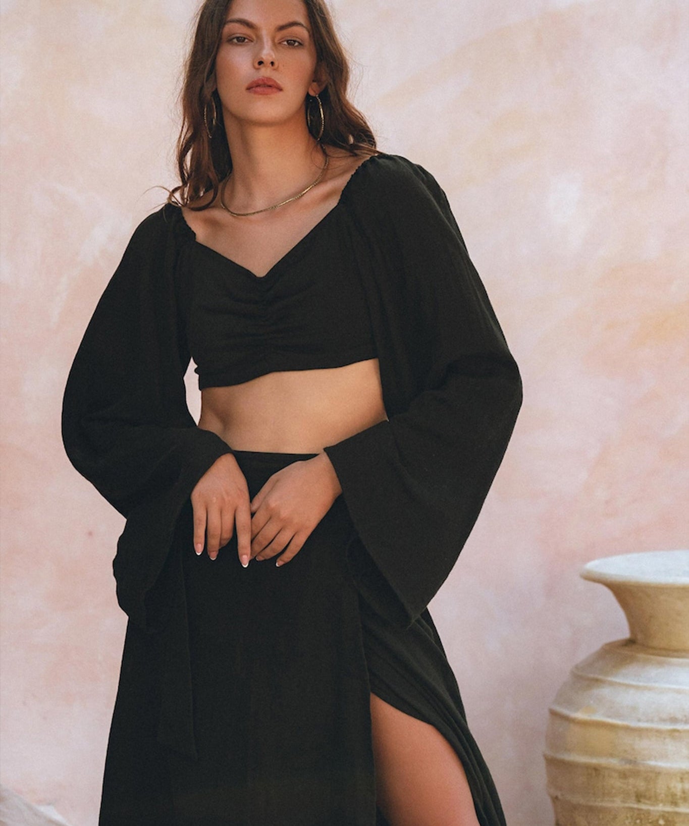 A person wearing the Black Summer Open Belly Top and Skirt by AYA Sacred Wear poses against a soft pink textured background. They have long hair and accessorize with hoop earrings. A large ceramic pot is partially visible on the right.