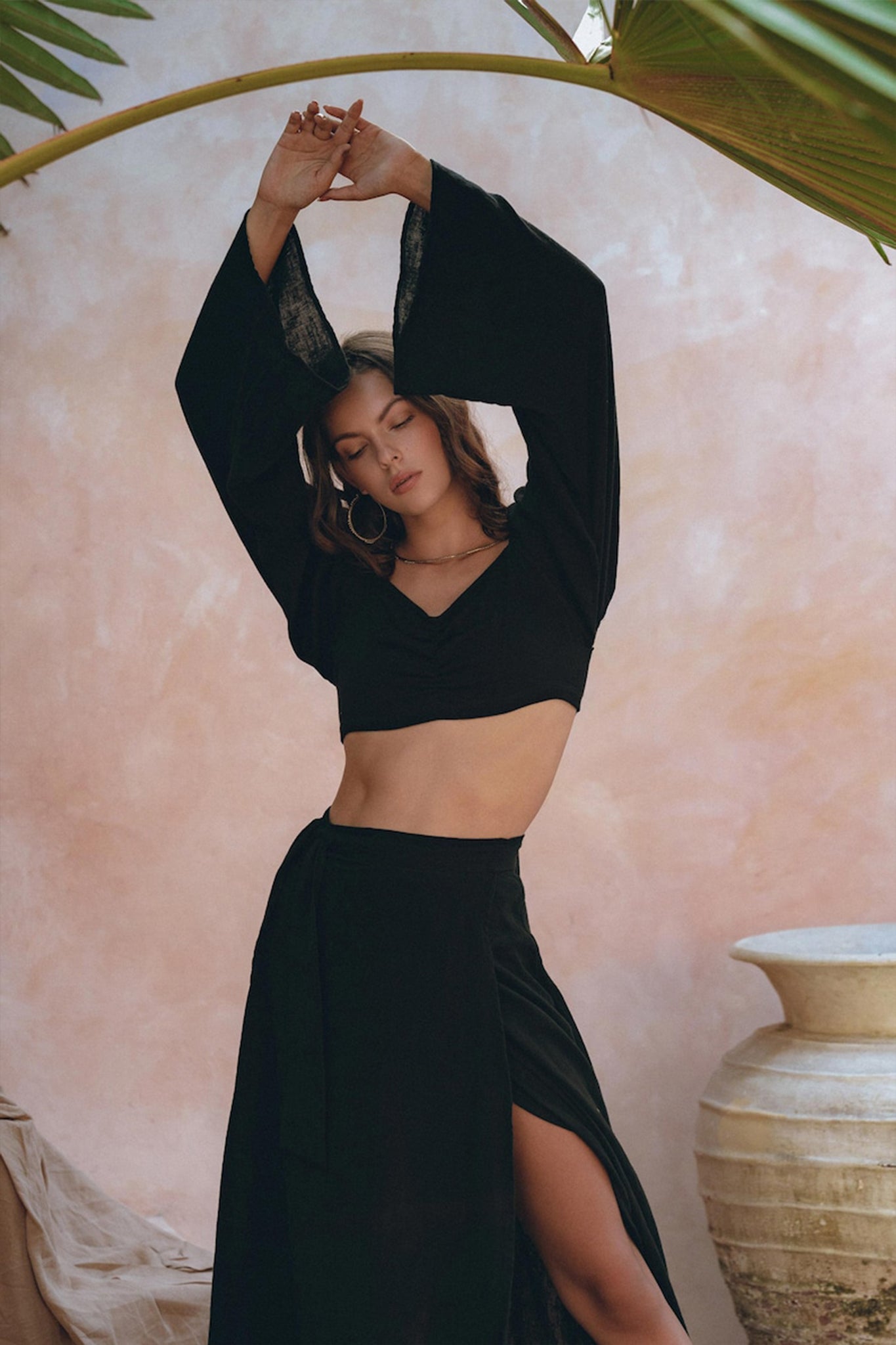 A woman in an elegant AYA Sacred Wear ensemble, featuring the Black Summer Open Belly Top and Skirt, poses gracefully with her arms raised against a textured pastel background. She stands next to a large decorative vase, surrounded by tropical leaves, exuding elegance and style.