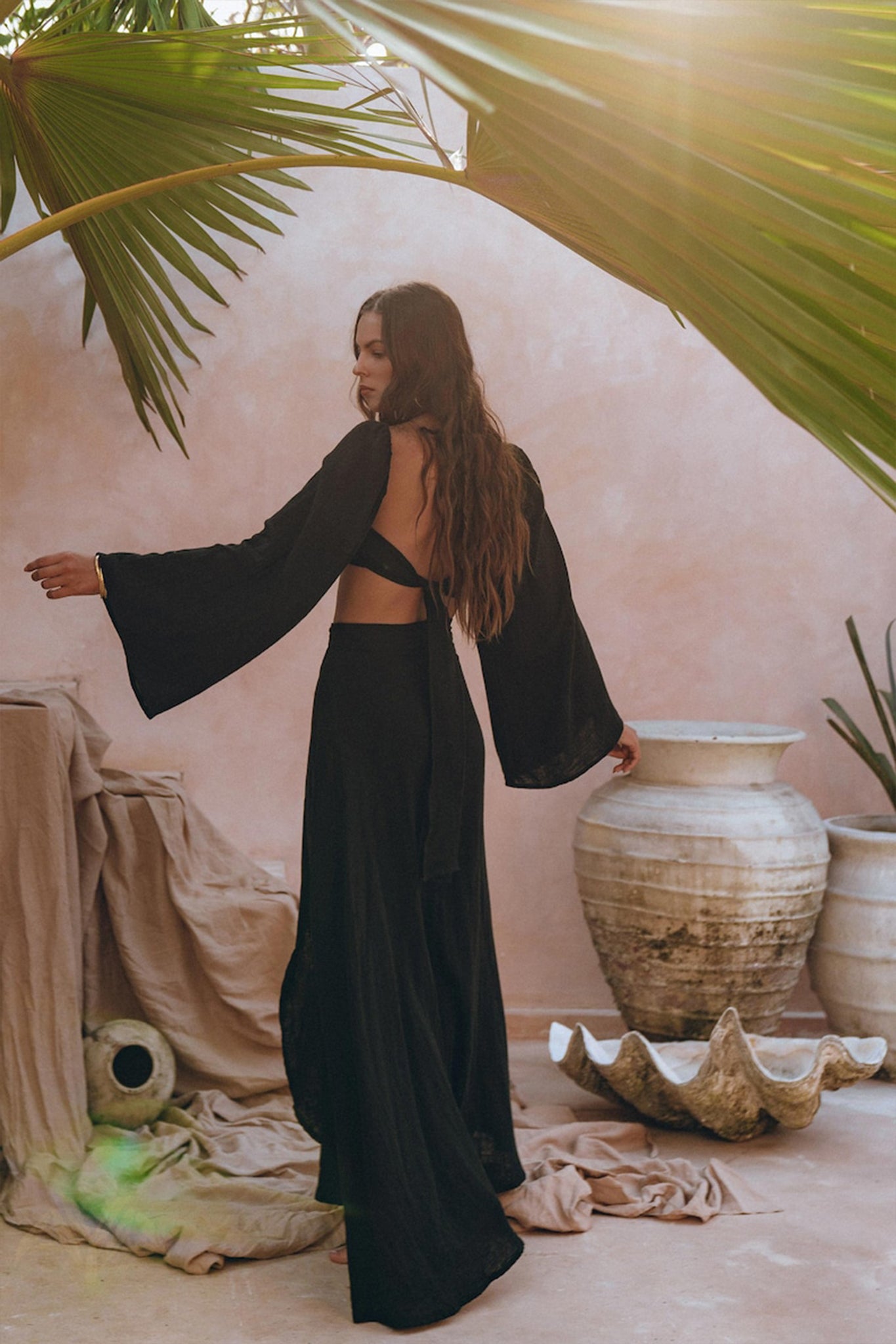 A woman with long hair, wearing a Black Summer Open Belly Top and Maxi Skirt by AYA Sacred Wear, stands in a sunlit room surrounded by large ceramic pots, draped fabric, and tropical plants. Sunlight filters through overhead palm leaves, casting shadows reminiscent of the ensemble's bohemian elegance.