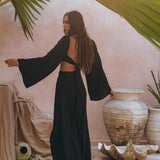 A woman with long hair, wearing a Black Summer Open Belly Top and Maxi Skirt by AYA Sacred Wear, stands in a sunlit room surrounded by large ceramic pots, draped fabric, and tropical plants. Sunlight filters through overhead palm leaves, casting shadows reminiscent of the ensemble's bohemian elegance.