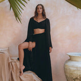 Wearing the Black Summer Open Belly Top and Skirt from AYA Sacred Wear, a woman poses gracefully in an elegant, tropical setting. She stands with one leg raised on a draped surface, surrounded by large clay pots and framed by green palm leaves against a textured peach wall.