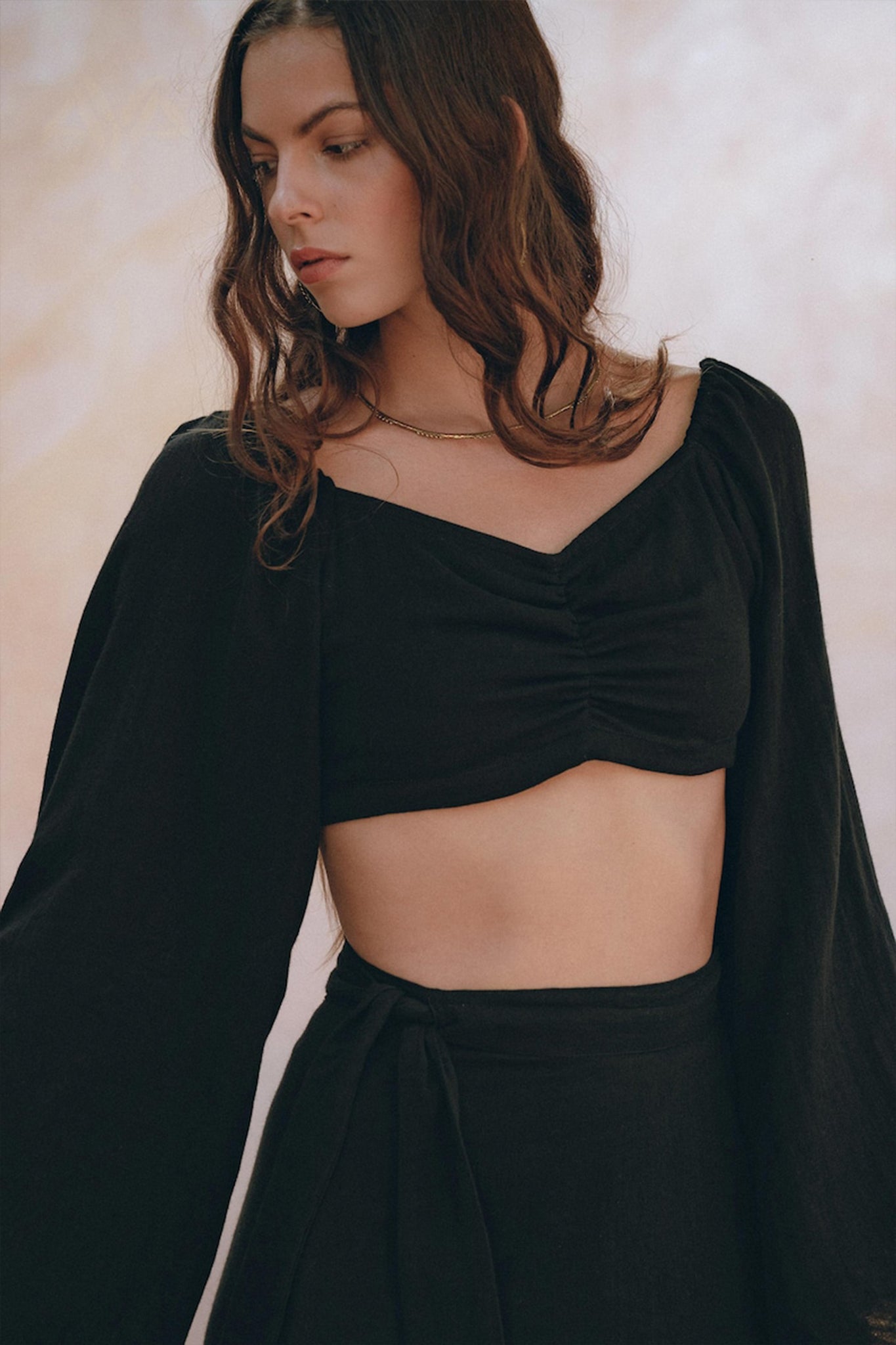 A woman with long brown hair is wearing an elegant Black Summer Open Belly Top and Skirt from AYA Sacred Wear, crafted with lightweight cotton fabric and featuring ruched detailing with long sleeves. She stands against a light, soft-focus background, looking down to her left with a serene expression.