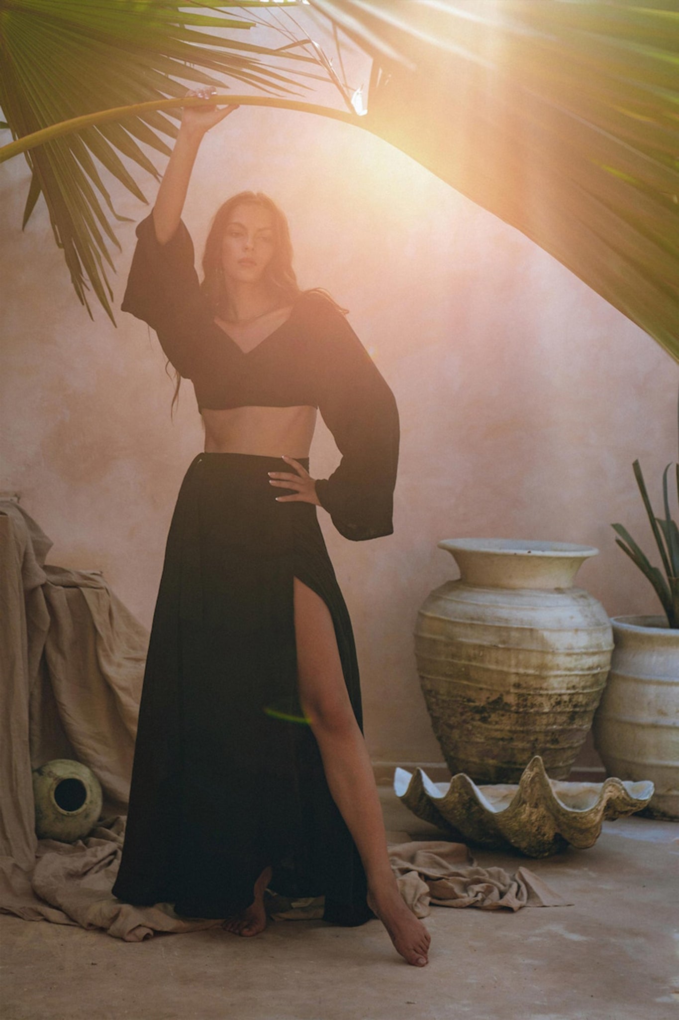 A woman models the Black Summer Open Belly Top and Skirt by AYA Sacred Wear, posing gracefully under a tropical palm frond. She stands barefoot on a sandy floor, encircled by large pottery and a shell-shaped sculpture. Sunlight filters through the lightweight cotton fabric of her black tank top and maxi skirt, creating a warm, serene atmosphere.