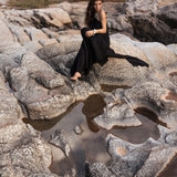 A woman wearing an AYA Sacred Wear Black Aurora Greek Goddess Dress, also known as a Cold Shoulder Maxi Dress or Gothic Grecian Dress, sits on rocky terrain near small pools of water. Her long hair flows freely as she gazes thoughtfully into the distance, with her arms lightly wrapped around her knees. The setting appears rugged and natural.
