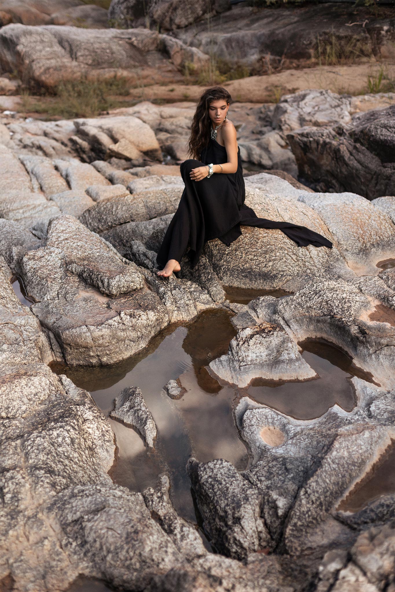 A woman wearing an AYA Sacred Wear Black Aurora Greek Goddess Dress, also known as a Cold Shoulder Maxi Dress or Gothic Grecian Dress, sits on rocky terrain near small pools of water. Her long hair flows freely as she gazes thoughtfully into the distance, with her arms lightly wrapped around her knees. The setting appears rugged and natural.