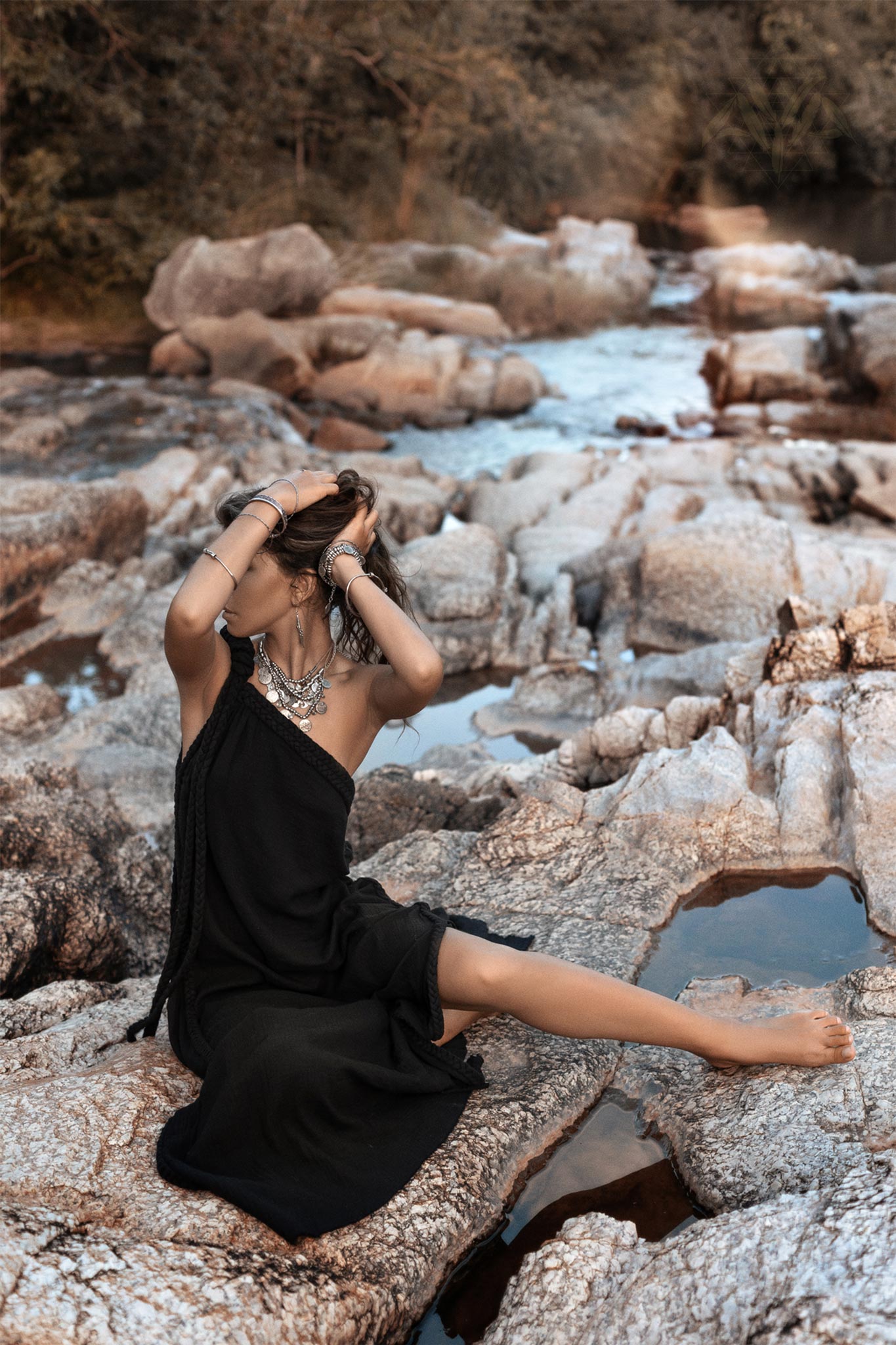 A woman in an AYA Sacred Wear Black Aurora Greek Goddess Dress • Cold Shoulder Maxi Dress • Gothic Grecian Dress sits barefoot on rocky terrain near a flowing river. She is adorned with several bracelets and a large necklace. One arm is raised to adjust her hair, and her other arm rests on the ground. The background features trees and water.