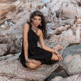 A woman wearing the Black Aurora Greek Goddess Dress by AYA Sacred Wear, crafted from organic cotton, sits on rocky terrain near a small body of water. She has long, wavy brown hair and is adorned with multiple pieces of jewelry. She looks directly at the camera with a calm expression while one leg is crossed over the other.