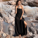 A woman wearing a long, Cold Shoulder Black Aurora Greek Goddess Maxi Dress by AYA Sacred Wear, adorned with various pieces of jewelry, stands barefoot on a rocky terrain. She gazes thoughtfully into the distance, her hand resting on her face, as the slit in her Gothic Grecian dress reveals part of her leg. The background features rugged rocks.