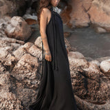 A person with long, wavy hair stands barefoot on rocky terrain near water. They are wearing the Black Aurora Greek Goddess Dress by AYA Sacred Wear, a flowing, sleeveless black dress with a Gothic Grecian style, along with assorted bracelets. The background consists of rough rocks and sparse vegetation.