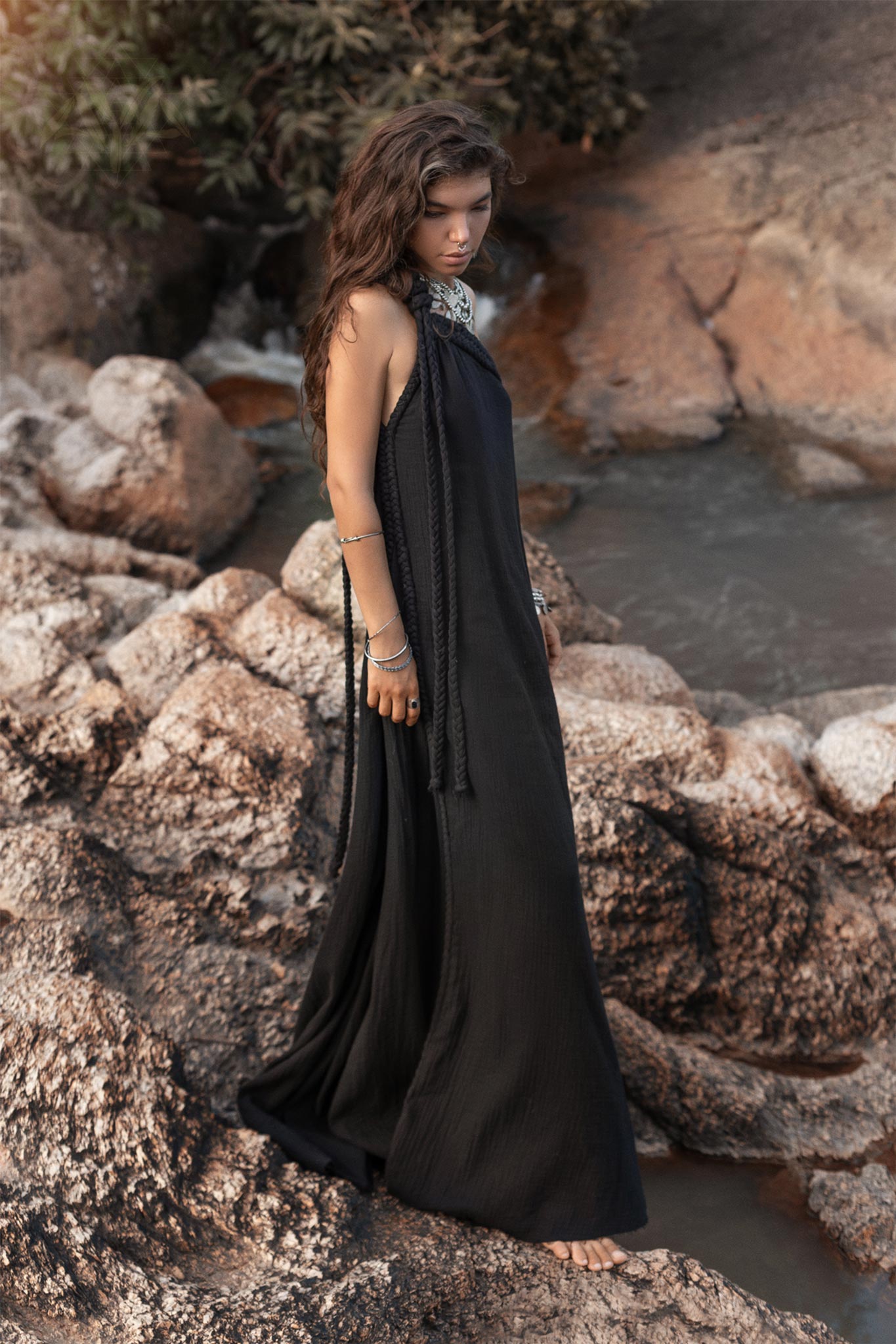 A person with long, wavy hair stands barefoot on rocky terrain near water. They are wearing the Black Aurora Greek Goddess Dress by AYA Sacred Wear, a flowing, sleeveless black dress with a Gothic Grecian style, along with assorted bracelets. The background consists of rough rocks and sparse vegetation.