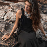 A woman with long, wavy hair sits on a rocky surface, wearing the Black Aurora Greek Goddess Dress by AYA Sacred Wear. The Cold Shoulder Maxi Dress and multiple layered necklaces complement her look. With a contemplative expression, she gazes to the side against a backdrop featuring rough, textured rocks.