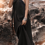 A woman stands on rocky terrain, wearing a cold shoulder Black Aurora Greek Goddess Dress by AYA Sacred Wear. She accessorizes with statement jewelry, including necklaces, bracelets, and rings, exuding a boho-chic aesthetic. The background features rugged rocks and a warm, glowing light.