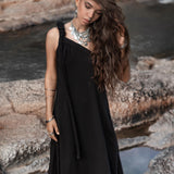 A woman with long, wavy hair and multiple necklaces stands outdoors on rocky terrain by the water. She is wearing a Black Aurora Greek Goddess Dress from AYA Sacred Wear, a cold shoulder maxi dress made of organic double-layer cotton. Her head is slightly tilted as she looks down with a serene expression, and her hand gently touches her hair.