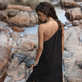 A woman with long wavy hair stands on rocky terrain near a stream, looking down. She is wearing a Black Aurora Greek Goddess Dress by AYA Sacred Wear and several bracelets. The background features rocks, water, and natural surroundings.