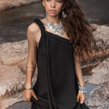 A person stands outdoors by a rocky waterway, wearing an AYA Sacred Wear Black Aurora Greek Goddess Dress • Cold Shoulder Maxi Dress • Gothic Grecian Dress adorned with braided detailing. They have long wavy hair and are accessorized with silver jewelry including necklaces, bracelets, and a nose ring. Their expression is neutral, and the setting appears natural and serene.